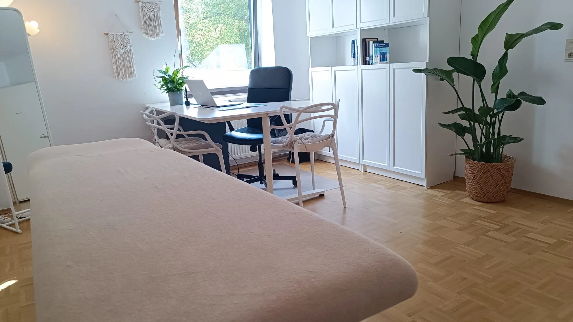 Heller Behandlungsraum mit Parkettboden: Behandlungsliege im Vordergrund, Schreibtisch am Fenster, große Zimmerpflanze.